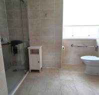 A bathroom with a walk-in shower, wall-mounted toilet with grab bars, small white cabinet, and beige tiled walls and floor.