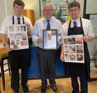 Three men in dress shirts and aprons stand indoors, holding up posters with multiple baby photos and a framed document, posing for a group photo.