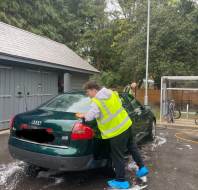 A person in a high-visibility vest and blue shoe covers washes the rear of a green Audi sedan outdoors near a building and bicycles.