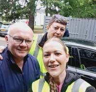 Three people wearing yellow safety vests stand in front of a black car, smiling for a selfie outdoors with trees and a fence in the background.
