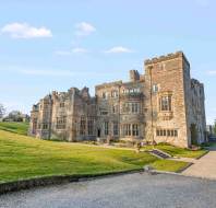A large stone castle with towers and many windows stands on a grassy hill, with a gravel driveway leading to the entrance under a clear sky.