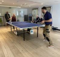 Two people play table tennis indoors while three others sit and watch; one person stands near the door in the background.