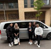 Five people stand smiling in front of a parked silver van on a city street, with modern apartment buildings and greenery in the background.