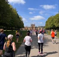 A group of people are running along a tree-lined path toward a large historic building on a sunny day.