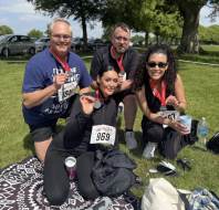 Four people with race bibs and medals sit on a blanket in a park, smiling at the camera, with trees, grass, and parked cars in the background.