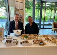 Two people sit behind a table displaying assorted baked goods and pastries for sale in a bright room with large windows showing greenery outside.