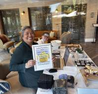 Two women sit at a table with baked goods, a laptop, and donation boxes. One woman smiles and holds a sign promoting a cake bake sale for a hospice fundraiser.