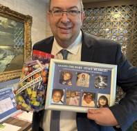 A man in a suit holds a jar of assorted sweets and a framed “Guess the Baby - Staff Members” photo collage featuring baby pictures of six individuals.