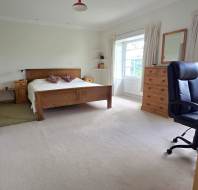 A tidy bedroom with a wooden bed, two dressers, a desk with an office chair, two windows, and white walls.