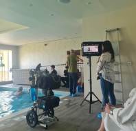 Film crew records a scene by an indoor pool. A camera operator films a swimmer, while others manage equipment and a woman watches the monitor.