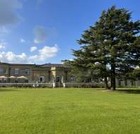 A large historic building with stone columns sits behind a spacious green lawn and a tall evergreen tree under a blue sky with scattered clouds.