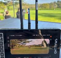 A camera monitor displays a live outdoor scene of people standing on grass, with three antennas attached on top of the monitor. Trees and a blue sky are visible in the background.