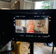 A monitor displays a film scene showing an older woman holding a red drink, standing in front of a bar, with camera equipment and a decorated room in the background.