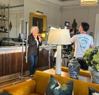 A woman stands near a bar holding a red cup while a cameraman films her in a well-decorated room with a lamp, couch, and framed portrait in the background.