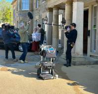 A film crew records two people standing outside a building entrance; cameras, boom mic, and equipment cart are visible in the foreground.