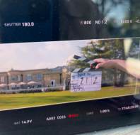 A film slate marked "212" is held in front of a large building and lawn, as seen on a camera monitor display during a film shoot.