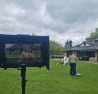 A camera monitor displays a filming scene on a lawn outside a building, with crew members and equipment visible in the background.