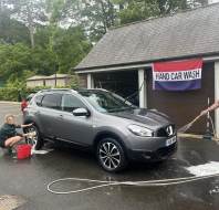Two people wash a gray SUV outside a building with a "Hand Car Wash" sign. One person scrubs the wheel while the other sprays water on the car.