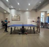 Two women are playing table tennis in a well-lit room while several people sit and watch from the side.