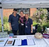 Three women stand behind a table with forms, raffle items, and cakes under a gazebo, likely at an outdoor fundraising or community event.