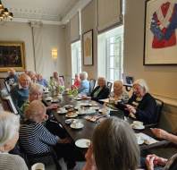 A group of elderly people sit around a long table having tea and snacks in a well-lit room with large windows and framed artwork on the walls.