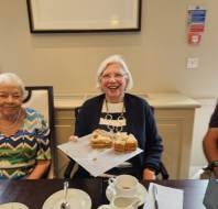 An older woman sits at a table holding a plate with slices of cake, smiling at the camera. Another woman sits beside her. There are cups, a jug, and papers on the table.