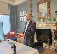 A man in a purple sequined jacket stands smiling behind a table with bottles and papers in an elegant, ornately decorated room.