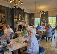 A group of elderly people sit at tables in a well-lit, elegant room with chandeliers, paintings, and large windows, appearing to participate in an activity or meeting.
