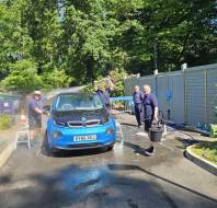 Four people wash a blue BMW i3 on a sunny day in a residential area, with cleaning equipment and water visible on the ground.