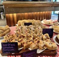 Various types of focaccia bread, including ones with sun-dried tomatoes, anchovies, and capers, are displayed on a checkered tablecloth with handwritten labels.
