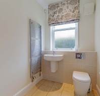 Small bathroom with a wall-mounted sink, toilet, heated towel rail, frosted window, patterned Roman shade, tiled floor, and a toilet paper holder.