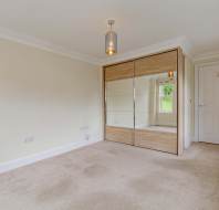Empty bedroom with beige carpet, cream walls, built-in mirrored wardrobe, ceiling light, and a window letting in natural light.