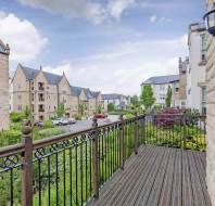 View from a balcony overlooking a landscaped garden, residential street, and stone buildings under a partly cloudy sky.
