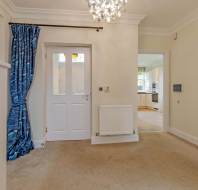 A hallway with beige carpet, a white door with glass panes, a blue curtain, and a view into a kitchen with wooden flooring and white cabinets.