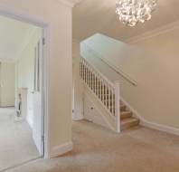 A hallway with carpeted stairs, cream walls, and a modern light fixture, leading to a living room with windows and beige carpeting visible through an open door.