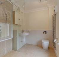 Modern bathroom with beige tiles, a glass-enclosed shower, a wall-mounted toilet, a small sink, a wall mirror, and a window with a patterned Roman shade.