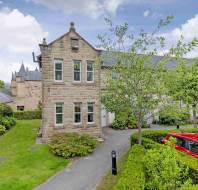 A stone residential building with two floors, surrounded by green lawns and trees, with a red car parked in the foreground on a paved driveway.