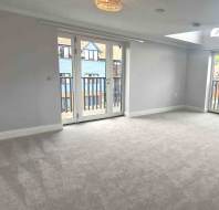 Empty room with light grey carpet, white walls, and two sets of glass doors leading to balconies with railings; natural light fills the space.