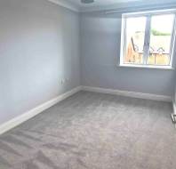 Small, empty room with light gray carpet, white walls, and a window facing out to neighboring buildings. Door is open, letting in natural light.