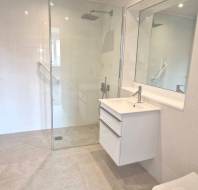 Modern bathroom with a walk-in shower, wall-mounted sink with drawers, large mirror, and wall-mounted toilet, all in a white and beige color scheme.