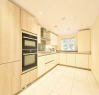 Modern kitchen with light wood cabinets, built-in oven, stovetop, neutral countertops, and a window letting in natural light. The floor is tiled in a light color.