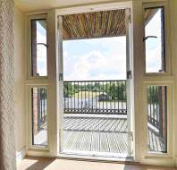 Glass double doors open onto a small balcony with a railing, overlooking a bright outdoor scene with a river, grassy areas, and a building in the distance.
