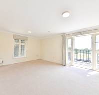 Empty, bright carpeted room with cream walls, large window, and double doors opening to a balcony with a view of greenery outside.
