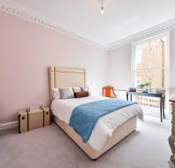 A neatly arranged bedroom with a double bed, light walls, crown molding, wooden furniture, and a large window letting in natural light.
