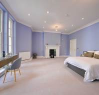 Spacious bedroom with light purple walls, a large bed, a desk and chair by tall windows, a white fireplace, and recessed ceiling lights.