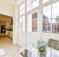 Bright modern kitchen and dining area with large windows, a round glass table, upholstered chairs, and a potted plant; view of buildings and trees outside.
