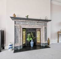 A decorative fireplace with a marble mantel, gold accent panels, and a blue vase with foliage in the center, surrounded by various ornaments on the hearth and nearby tables.