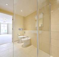 Modern bathroom with beige tiles, glass shower enclosure, wall-mounted toilet and bidet, large mirror, and view into a bright adjoining room with windows.