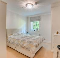 A tidy bedroom with a double bed, patterned bedding, a small window with a roman shade, a white dresser, a stool, and overhead lighting.