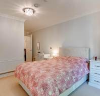 A bedroom with a double bed covered in a pink patterned duvet, white bedside tables, a white dresser, and neutral walls, lit by a ceiling light.